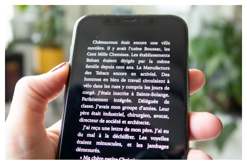 Une personne tient un smartphone dans sa main. Le plan se concentre sur le contenu du téléphone : un extrait de livre. L'ebook est lu sur fond noir, avec texte blanc. En arrière plan se trouvent des plantes, illuminée par un beau soleil d'été.